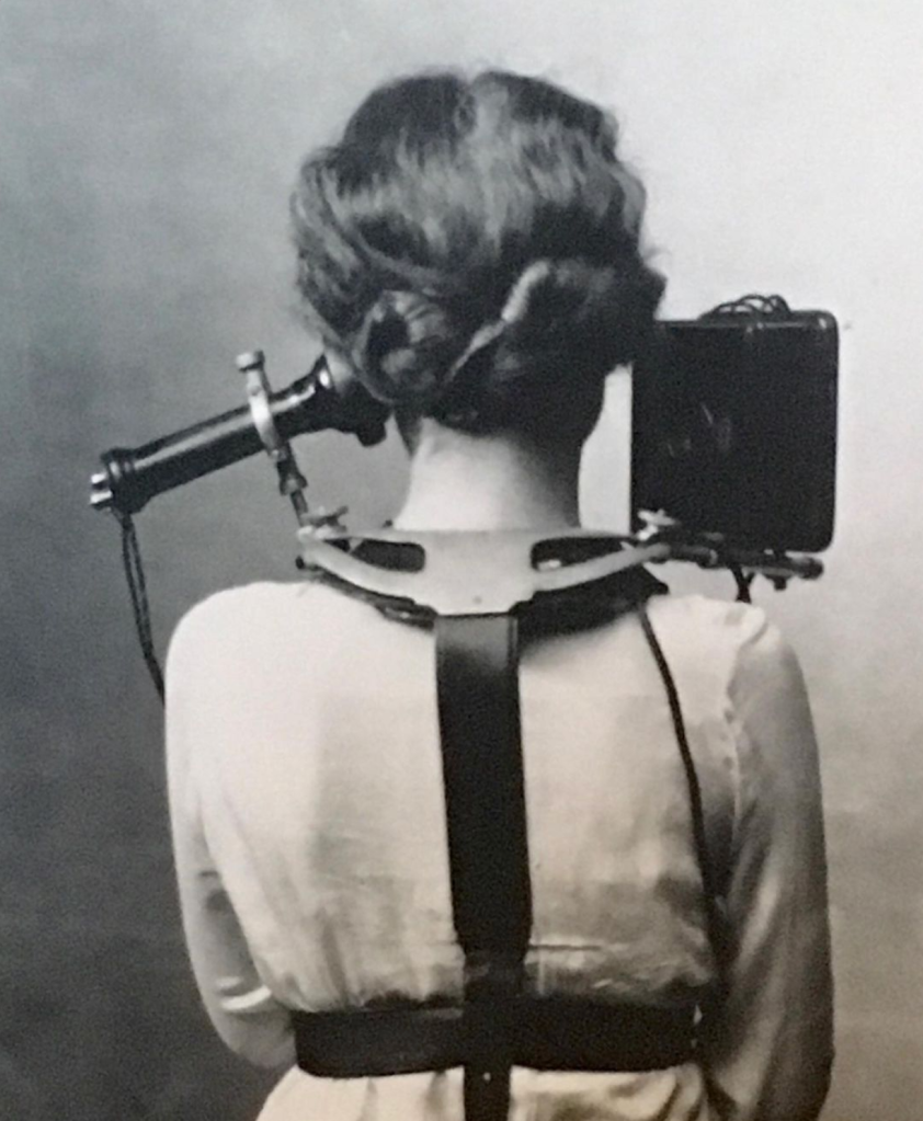 Black and white photo of a woman from behind, wearing a back brace that supports an old-style phone earpiece beside one ear and a mouthpiece beside the other for "hands free" phone use. 